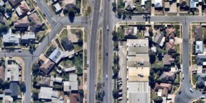 Vista aérea de um bairro suburbano bem planejado, com uma avenida principal dividida por canteiro central, cercada por casas residenciais com quintais e garagens. À direita, há edifícios comerciais maiores com estacionamentos e veículos visíveis. O tráfego está ativo nas ruas, e há árvores espalhadas pela área.