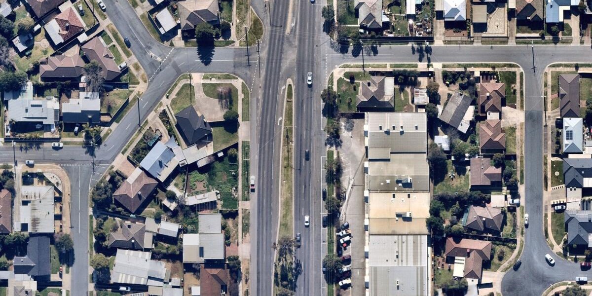 Vista aérea de um bairro suburbano bem planejado, com uma avenida principal dividida por canteiro central, cercada por casas residenciais com quintais e garagens. À direita, há edifícios comerciais maiores com estacionamentos e veículos visíveis. O tráfego está ativo nas ruas, e há árvores espalhadas pela área.