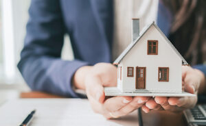 Pessoa vestida com terno segurando uma miniatura detalhada de uma casa sobre a mesa, com documentos e uma caneta ao fundo, sugerindo contexto profissional relacionado a imóveis ou planejamento financeiro.