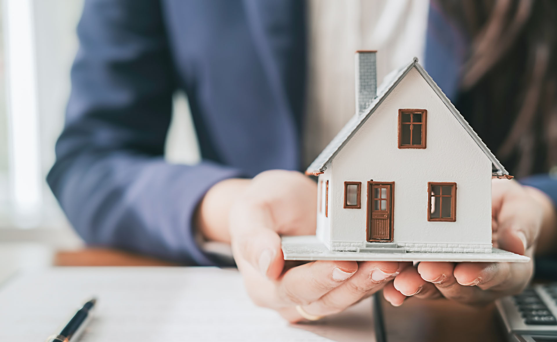 Pessoa vestida com terno segurando uma miniatura detalhada de uma casa sobre a mesa, com documentos e uma caneta ao fundo, sugerindo contexto profissional relacionado a imóveis ou planejamento financeiro.