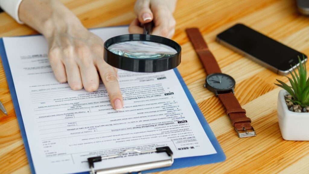 Pessoa fiscalizando documentos com lupa, sugerindo a busca por omissão de receita ou irregularidades financeiras. Um relógio e um telefone estão na mesa.